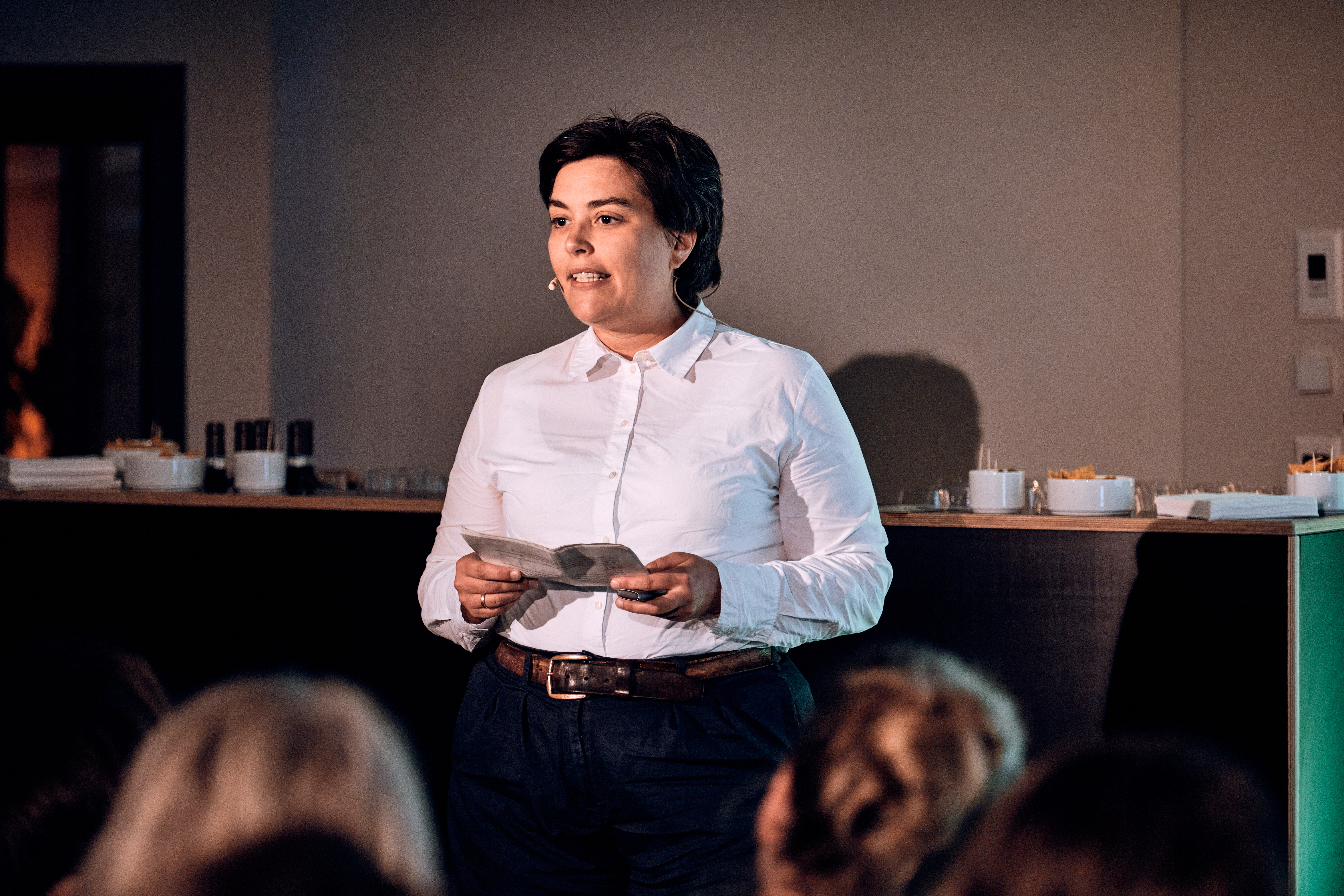 Eine Person in einem weißen Hemd mit Knöpfen steht vor einem Publikum, spricht und hält Papiere, während im Hintergrund Erfrischungen gereicht werden.